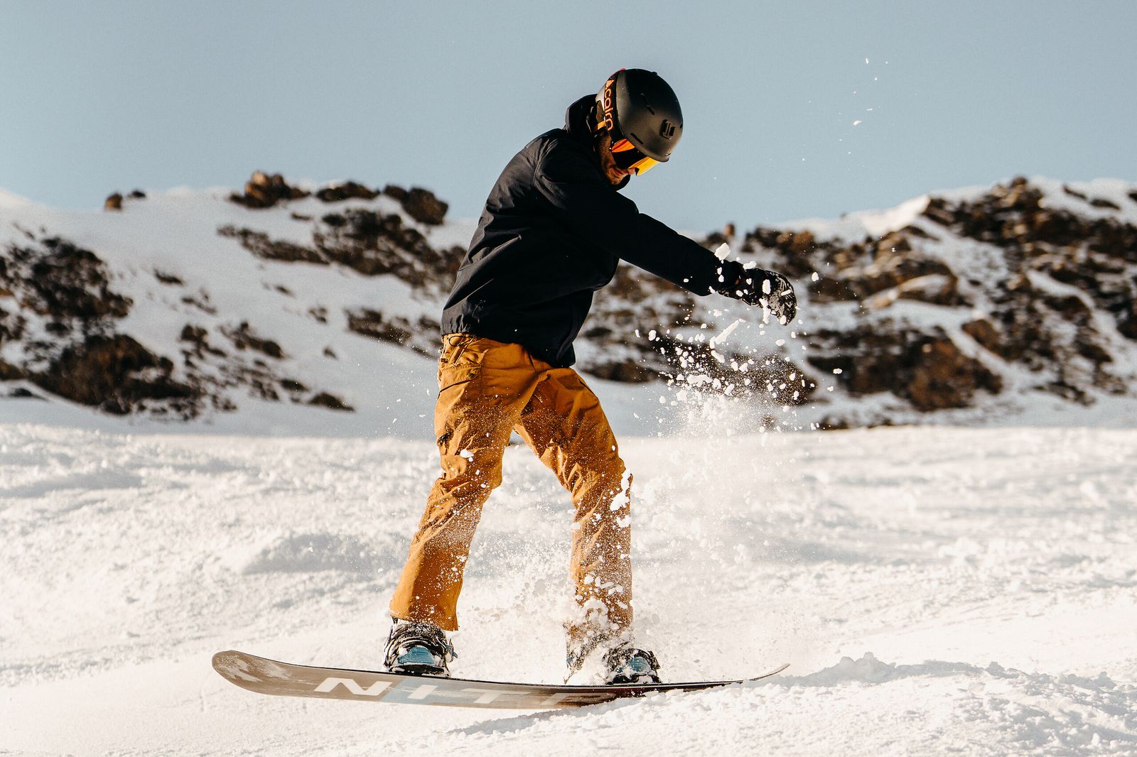 Snowboarder fazendo curva e levantando neve, com montanhas rochosas ao fundo